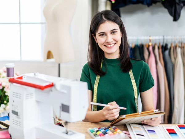 Smiling caucasian female fashion designer works in studio by idea drawing sketches with digital tablet and colorful fabric for a dress design collection, choose clothing colors for tailoring